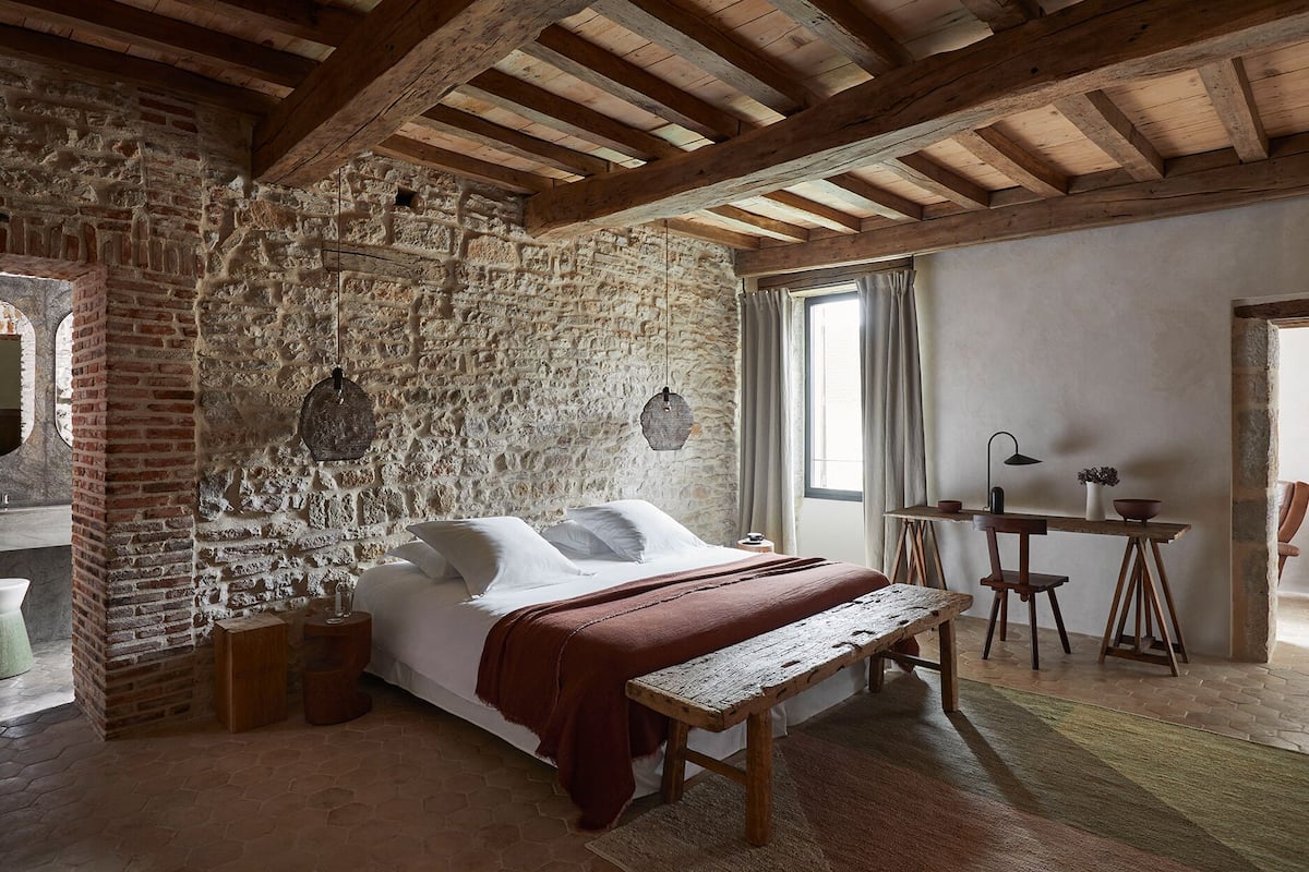 A rustic bedroom with exposed stone walls, a wooden bed, a bench, and a small table with a lamp.