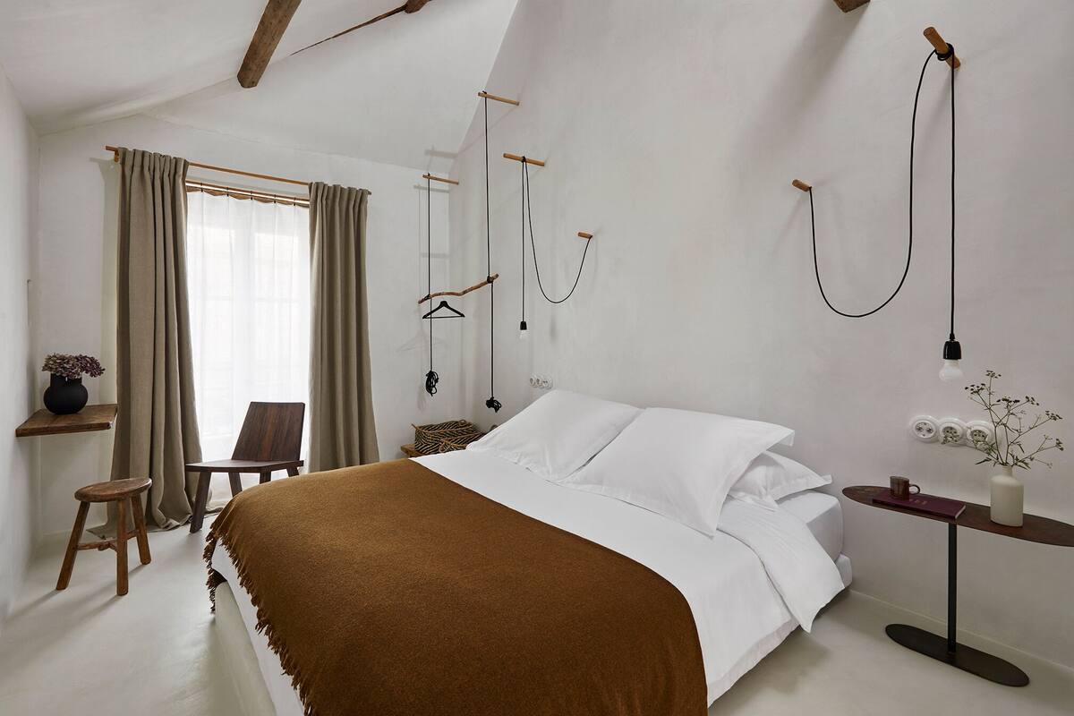 A bedroom with a large bed, a wooden chair, a small table with a vase, and a hanging light fixture.