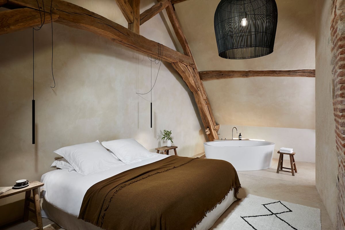 A bedroom with a large bed, a wooden ceiling, a hanging light fixture, a bathtub, and a small stool.
