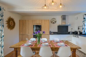 A dining area with a wooden table set for four, surrounded by white chairs. A vase with purple flowers is on the table.