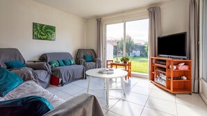 A living room with a sofa, a coffee table, a TV on a wooden stand, and a view of the outdoors through a large window. - La Massane - Three Bedroom House, Sleeps 6 (Argeles-sur-Mer)