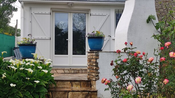 A house with a white exterior, a front door, and a stone staircase leading to the entrance. There are two large blue flower pots on the wall, and a variety of flowers and plants around the house. - L'Orchidée - Two Bedroom House, Sleeps 6 (Limerzel)