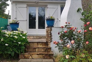 Une maison à l’extérieur blanc, avec une porte d’entrée et un escalier en pierre menant à l’entrée. Deux grands pots de fleurs bleus sont placés sur le mur, et une variété de fleurs et de plantes ornent les alentours de la maison.