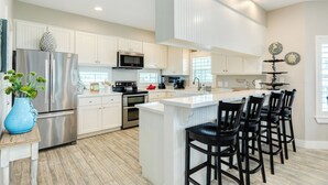 A modern kitchen with white cabinets, stainless steel appliances, and a central island with bar stools. - Spacious Beachside Home w/ Long Gulf View, Covered Deck & Huge Primary Suite (Galveston)