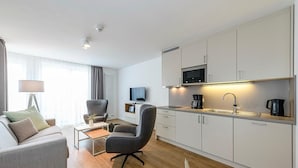 Ein modernes Wohnzimmer mit einer Couch, einem Sessel und einem Couchtisch. Ein Flachbildfernseher ist an der Wand montiert. Die Küche verfügt über weiße Schränke, eine Mikrowelle und eine Kaffeemaschine.