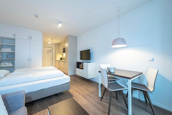 A modern hotel room with a bed, a desk, a chair, a television, and a wardrobe.