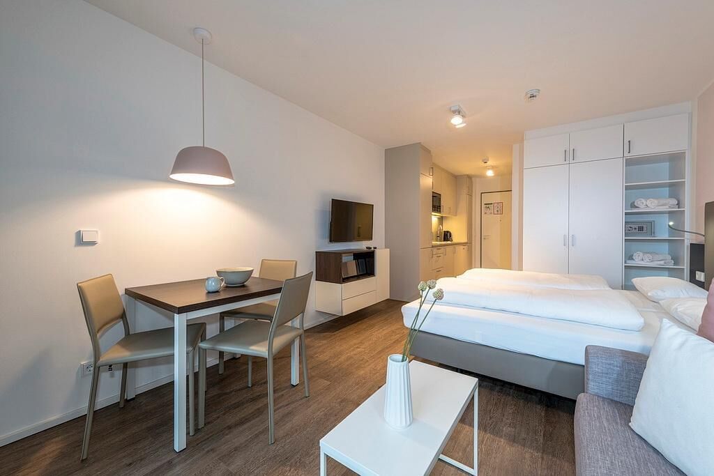 A modern hotel room with a bed, a dining table with chairs, a TV, and a kitchenette.
