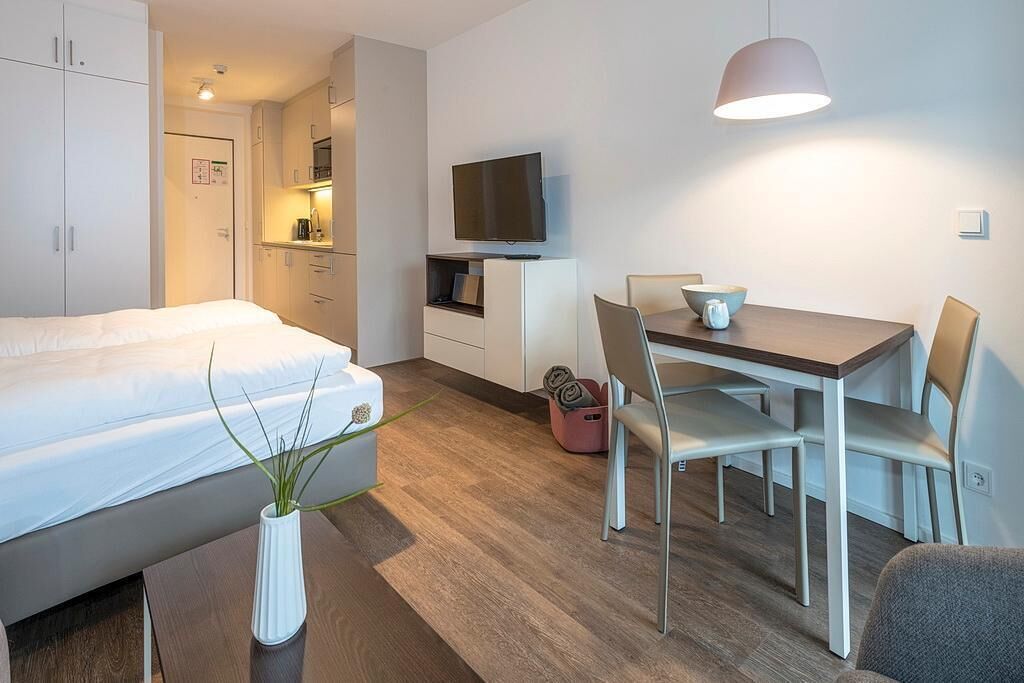 Een moderne hotelkamer met een bed, een eettafel met stoelen, een televisie en een klein keukentje.