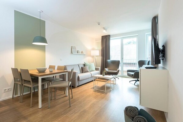 A modern living room with a dining area, a sofa, a coffee table, and a TV mounted on the wall. - Modern family apartment near the beach with sheltered loggia & sauna in the house! (Nordseeheilbad Langeoog)