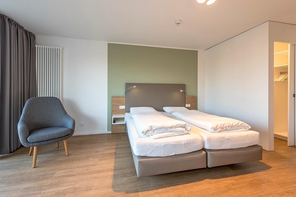 A modern hotel room with a double bed, a grey armchair, and a wooden floor.
