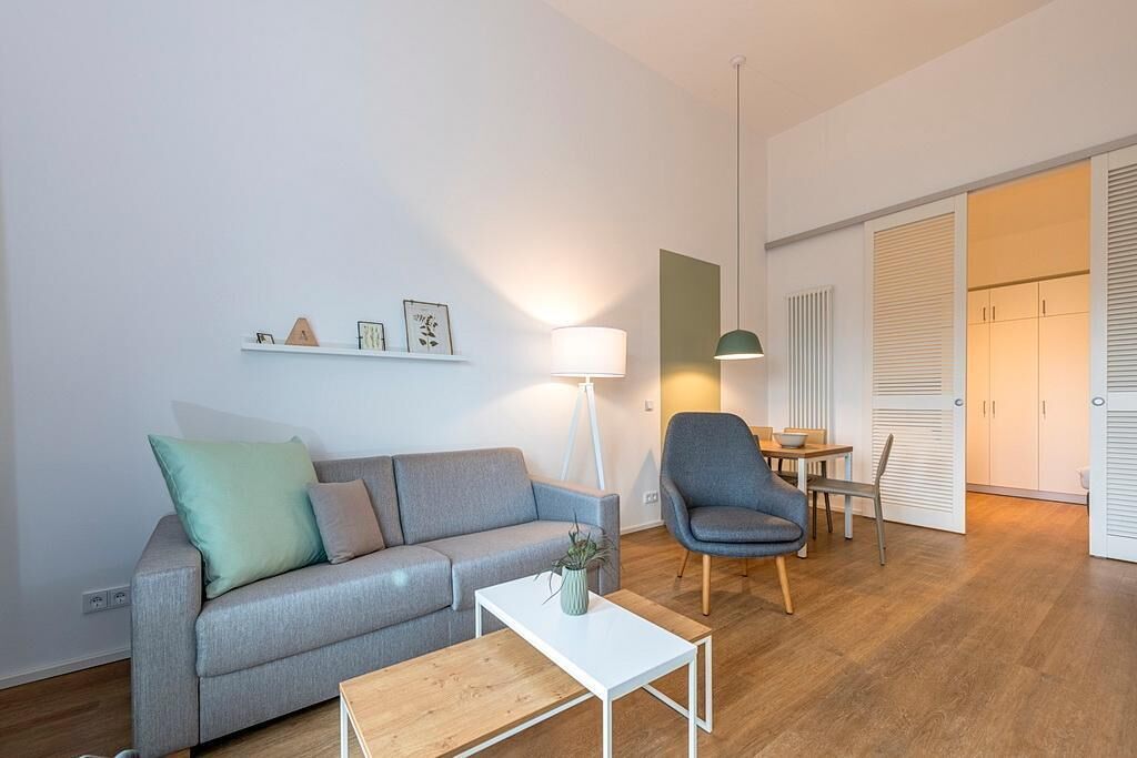 Ett modernt vardagsrum med en grå soffa, ett träbord, en blå fåtölj och en lampa. Det finns ett litet bord med en skål och en växt. Rummet har trägolv och vita väggar.