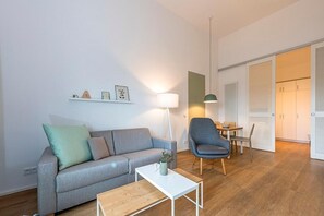 Una sala de estar moderna con un sofá gris, una mesa de centro de madera, un sillón azul y una lámpara. Hay una mesita con un cuenco y una planta. El piso es de madera y las paredes son blancas.