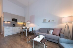 Una sala moderna con un sofá gris, una mesa de centro, un comedor con sillas y una cocina con armarios blancos y un microondas.