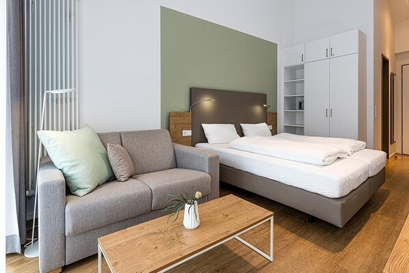 Et moderne hotelværelse med en grå sofa, et træbord, en seng med hvide sengetæpper og et hvidt skab.