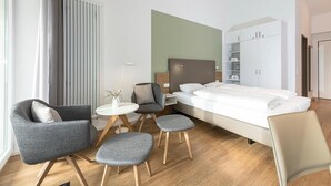 A modern hotel room with a bed, two armchairs, a small table, and a cabinet.