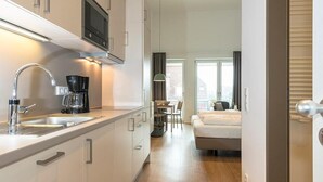 A modern kitchen with a built-in oven, a coffee maker, and a sink. There is a dining area with a table and chairs, and a bedroom with a bed and a window.