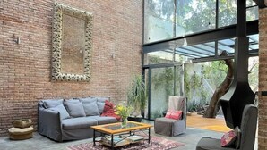 A modern living room with a brick wall, a grey sofa, a wooden coffee table, and a glass-enclosed patio area with a tree. - Luxury Stays (Tlacopac, Álvaro Obregón)