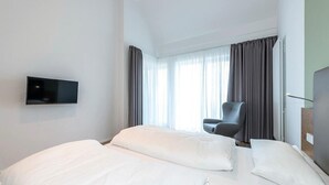A hotel room with a bed, a chair, a flat-screen TV mounted on the wall, and a window with curtains.