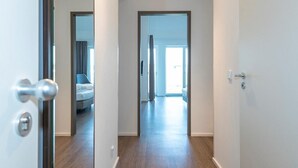 A hallway with a wooden floor and white walls, leading to a bedroom with a bed and a window.
