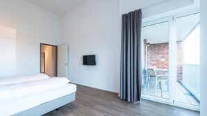 A modern hotel room with a bed, a flat-screen TV, a sliding glass door to a balcony with a brick wall and outdoor furniture.