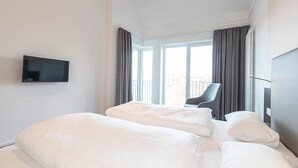 A hotel room with two beds, a TV mounted on the wall, a chair, and a balcony with curtains.