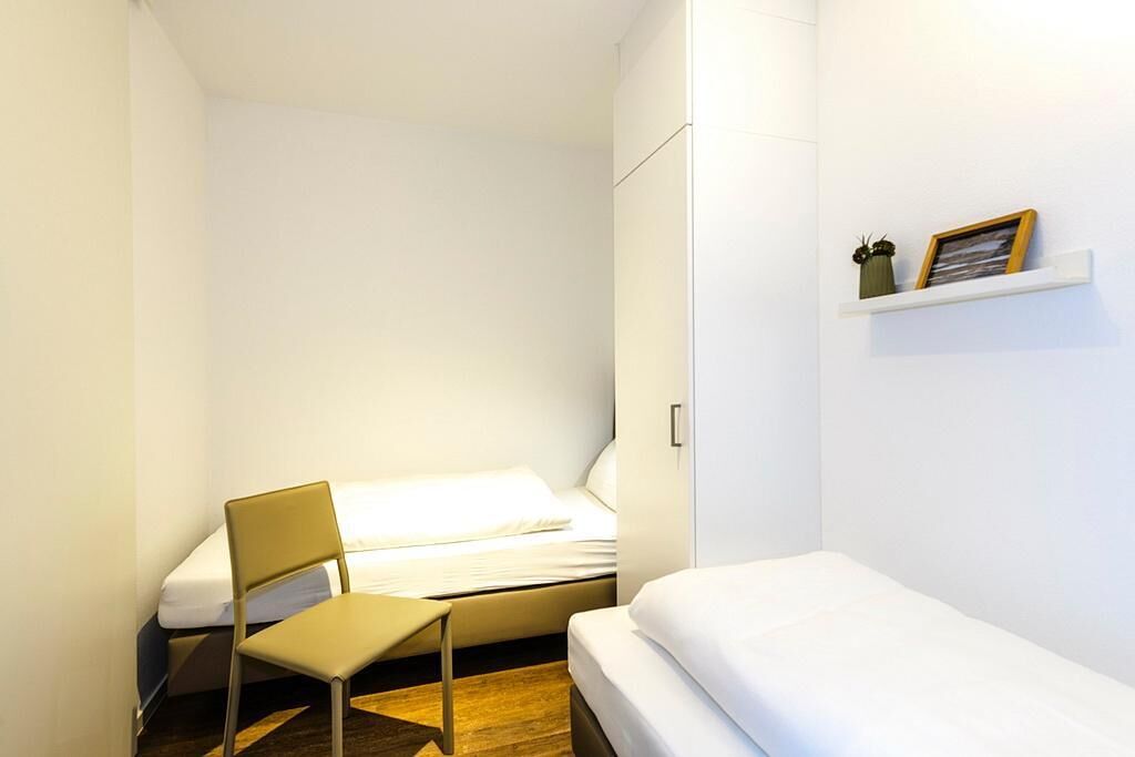 Un dormitorio pequeño y ordenado con una cama individual, una silla y una estantería con un cuadro y una planta.
