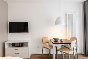 A modern hotel room with a flat-screen TV mounted on the wall, a wooden dining table with two chairs, a white bedside table with drawers, and a radiator with curtains.