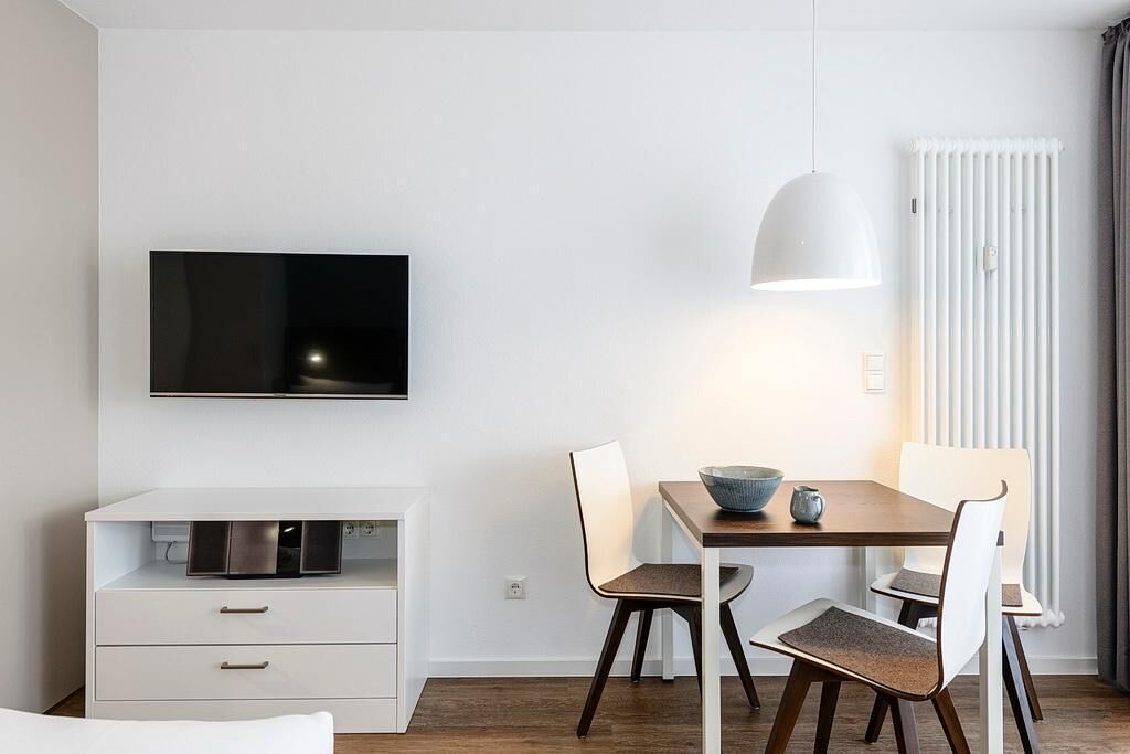 Een moderne, minimalistisch ingerichte kamer met een flatscreen televisie aan de muur, een eettafel met stoelen, een witte kast met lades en een radiator.