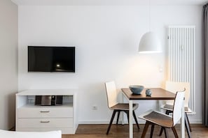 Una stanza moderna e minimalista con una TV a schermo piatto montata a parete, un tavolo da pranzo con sedie, un armadietto bianco con cassetti e un termosifone.