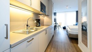 Cocina moderna con horno integrado, cafetera y lavamanos. Hay un comedor con mesa y sillas, además de un dormitorio con cama y ventana.