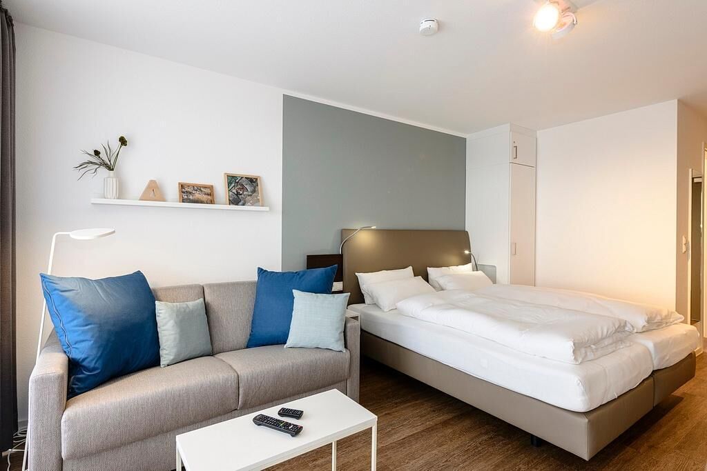 A modern bedroom with a sofa, a bed, a bedside table, and a small white shelf with decorative items.
