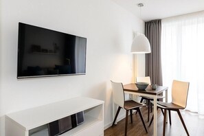 Ein modernes Hotelzimmer mit einem wandmontierten Flachbildfernseher, einem Esstisch mit Stühlen, einem weißen Konsolentisch und einem großen Fenster mit Vorhängen.