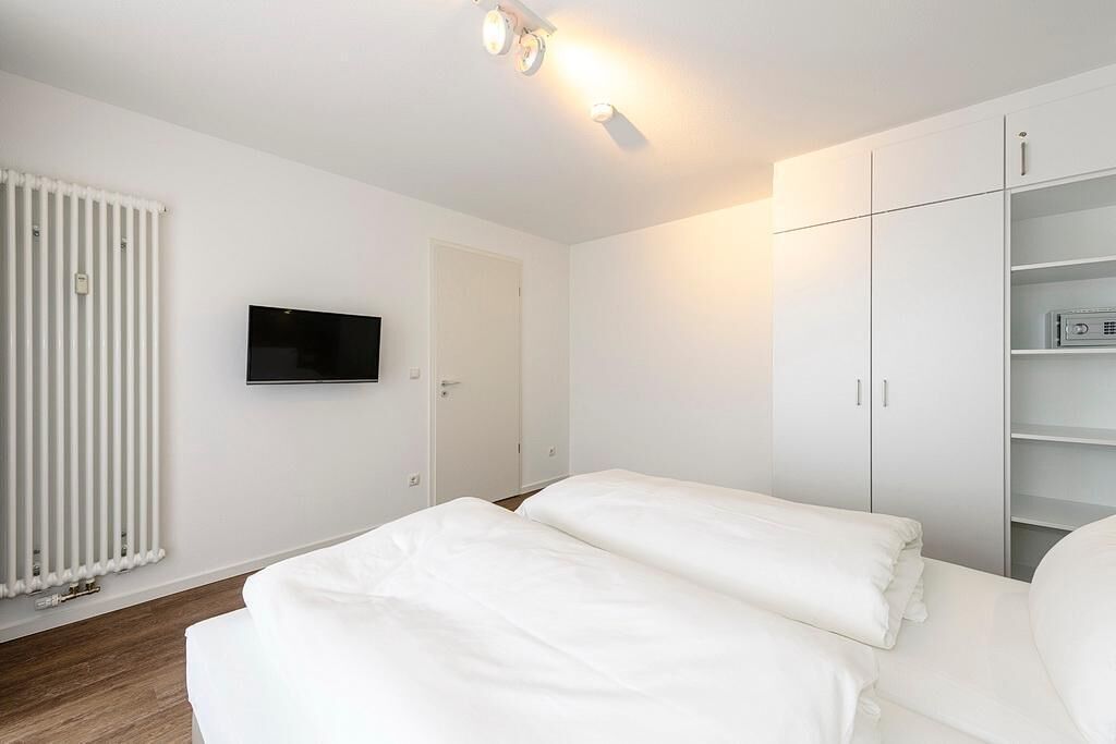 Ein modernes, minimalistisches Schlafzimmer mit einem großen Bett, einem an der Wand montierten Fernseher und einem Kleiderschrank mit offenen Regalen.