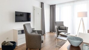 Ein modernes Wohnzimmer mit Flachbildfernseher, zwei grauen Sesseln, einem Couchtisch und einer Stehlampe.