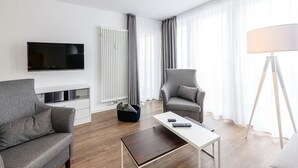 Ein modernes Hotelzimmer mit einem Flachbildfernseher, zwei grauen Sessel, einem weißen Couchtisch und einer Stehlampe.