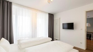 Ein modernes Hotelzimmer mit zwei weißen Betten, einem flachbildfernseher an der Wand und Blick ins Freie durch große Fenster mit durchsichtigen Vorhängen.