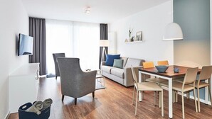 Una sala de estar moderna con un sofá, una mesa de comedor y sillas. Hay un televisor montado en la pared, una lámpara y un estante con adornos.