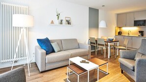 Una sala de estar moderna con un sofá, una mesa de centro y un comedor con sillas y una mesa. Hay una zona de cocina con armarios y electrodomésticos.