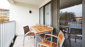 Un balcón exterior con mesa y sillas de madera, barandilla de metal y vistas de la ciudad.