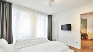 Habitación de hotel con una cama grande, un televisor de pantalla plana en la pared y vistas al exterior a través de cortinas translúcidas.