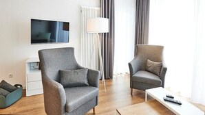 Una sala de estar moderna con dos sillones grises, una mesa de centro blanca, un televisor de pantalla plana, una lámpara de pie y un radiador.
