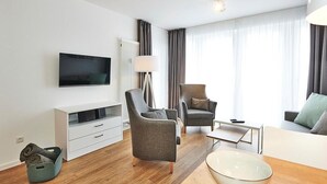 Un salón moderno con una televisión de pantalla plana, dos sillones grises, un sofá blanco, una mesa de centro y una lámpara de pie.