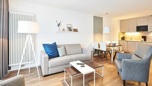 Ein modernes Wohnzimmer mit einer Couch, einem Couchtisch und einem Sessel. Es gibt einen Essbereich mit Tisch und Stühlen sowie eine Küchenzeile mit Schränken und Geräten.