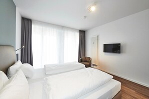 Habitación de hotel con dos camas, una silla, un televisor y un ventanal con cortinas.