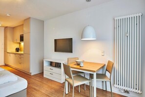 A modern kitchen with white cabinets, a dining table with chairs, a TV mounted on the wall, and a radiator. - Strandnahes 1-zimmer Ferienapartment mit Balkon Inkl. Schwimmbad- & Saunanutzung (Nordseeheilbad Bensersiel)
