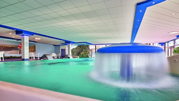 Una piscina interior con tobogán de agua, techo azul y blanco, y zonas de estar.
