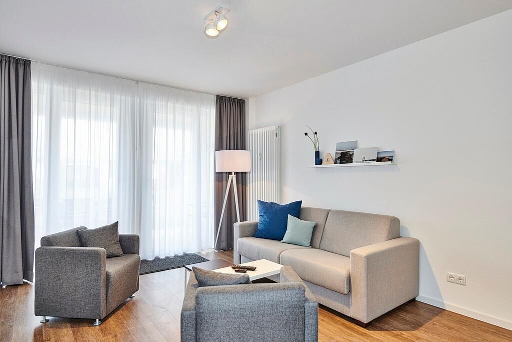 A modern living room with a sofa, armchair, and coffee table. There is a lamp, a radiator, and a shelf with decorative items.