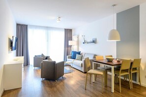 A modern living room with a sofa, chairs, a dining table, and a TV. - Familienapartment Nahe Nordsee mit Balkon Inkl. Schwimmbad- & Saunanutzung (Nordseeheilbad Bensersiel)
