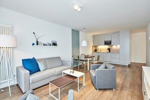 Una sala de estar moderna con un sofá gris, una mesa de centro y un sillón gris. Hay un comedor con una mesa y sillas, y una cocina con armarios blancos y un microondas.