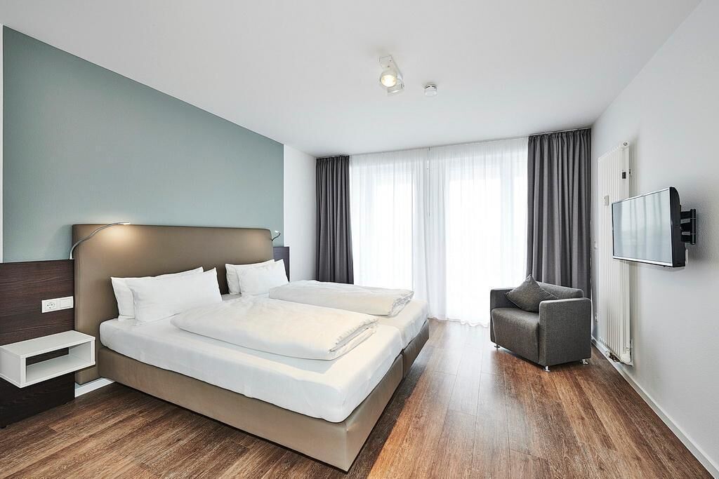 A modern hotel room with a large bed, a bedside table, a chair, a TV, and a window with curtains.